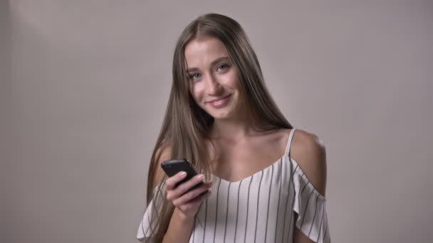 Молодая привлекательная девушка держит смартфон, смотрит на камеру, улыбается, концепция общения, серый фон — стоковое видео