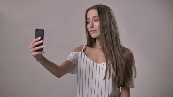 Joven chica tímida atractiva está haciendo selfie en el teléfono inteligente, fijación de cabello, concepto de comunicación, fondo gris — Vídeos de Stock