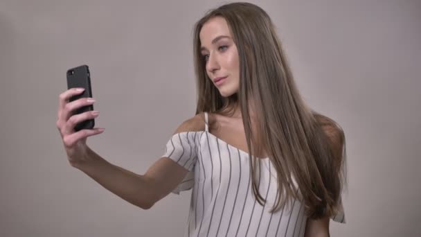 Joven chica tímida atractiva está haciendo selfie en el teléfono inteligente, concepto de comunicación, fondo gris — Vídeos de Stock