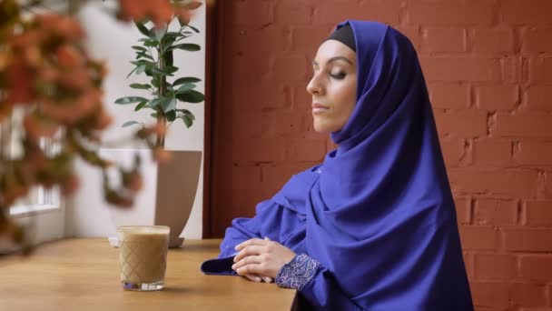 Молодая мусульманка в хиджабе сидит в кафе и смотрит в камеру, серьезная красивая женщина в платке — стоковое видео