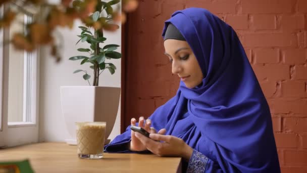 Молодая мусульманка в хиджабе, печатает по телефону и сидит в кафе, красивая женщина с проколотым носом — стоковое видео