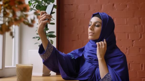 Wanita muslim muda dalam jilbab mengambil selfie dan tersenyum, duduk di kafe, wanita menawan dengan hidung ditindik — Stok Video