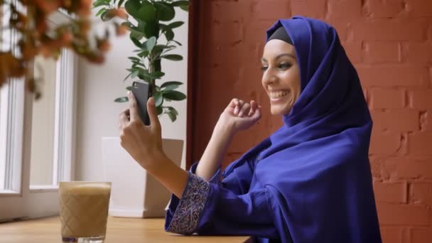 Jonge moslimvrouw in hijab video praatje via mobiele telefoon, lacht, zitten in moderne Cafe — Stockvideo