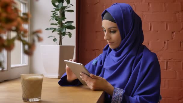 Wanita muslim muda yang cantik mengenakan jilbab biru menggunakan tablet dan duduk di kafe, wanita menawan dengan hidung yang ditindik — Stok Video