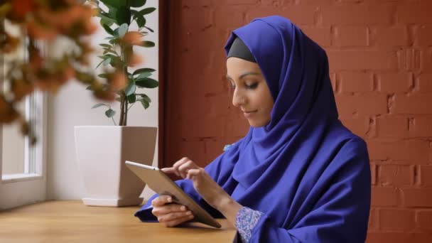Jonge moslimvrouw in blauwe hijab te typen op tablet en zitten in moderne Cafe, charmante dame met doordrongen neus — Stockvideo