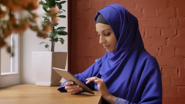 Wanita muslim muda yang menawan dengan jilbab biru mengetik di tablet dan tersenyum di depan kamera, duduk di kafe, wanita cantik dengan hidung yang ditindik — Stok Video