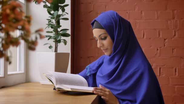 Jonge mooie Moslimvrouw in blauwe hijab lezen boek in café en glimlachend in de camera, charmante vrouw met doordrongen neus — Stockvideo