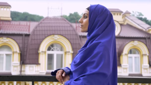 Profil av unga självsäkra muslimska kvinnan i hijab vänder och tittar på kameran, stående på balkong — Stockvideo