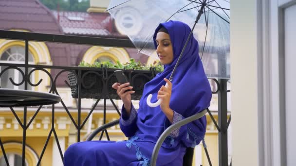Wanita muslim muda berhijab duduk di kursi di balkon dengan payung dan mengetik di ponsel — Stok Video