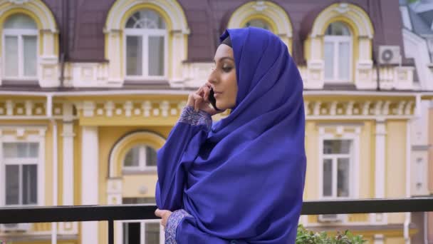 Jeune femme musulmane en hijab debout sur le balcon et parlant au téléphone, vue sur la rue — Video