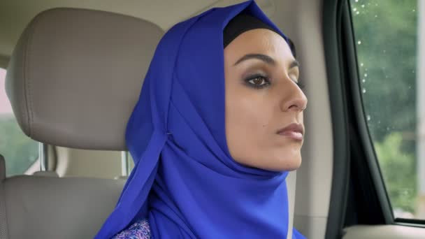 Joven mujer musulmana en hijab en coche en el asiento trasero del pasajero mirando hacia adelante, serio — Vídeos de Stock