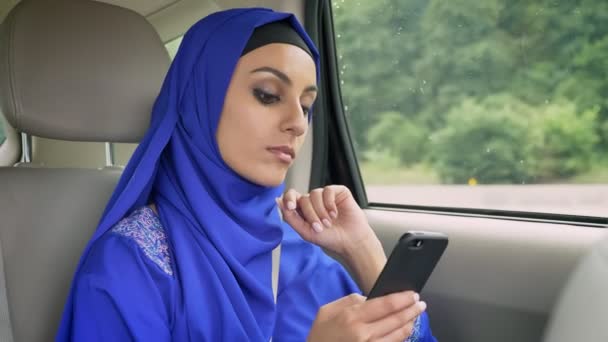 Junge schöne muslimische Frau im Hidschab im Auto auf dem Beifahrersitz tippt mit ernstem Gesichtsausdruck auf Handy — Stockvideo