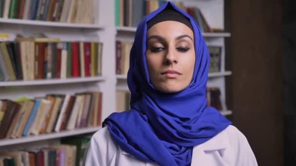 Jeune belle femme musulmane en hijab debout dans la bibliothèque et regardant la caméra avec une expression sérieuse — Video