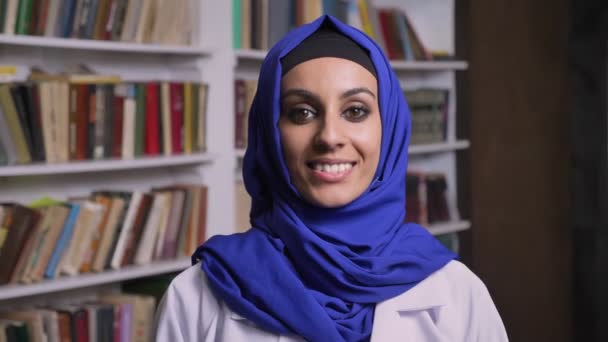 Junge schöne muslimische Frau im Hidschab steht in der Bibliothek und lächelt mit glücklichem Gesichtsausdruck in die Kamera — Stockvideo