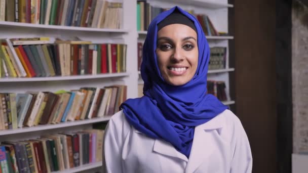 Junge glückliche muslimische Frau im Hidschab, die in der Bibliothek steht, in die Kamera schaut und lächelt — Stockvideo
