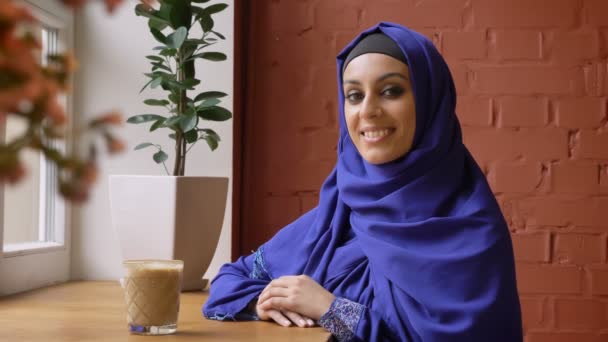 Unga muslimska kvinnan i hijab tittar på kameran och ler, sitter i moderna café, dam med piercade näsan — Stockvideo