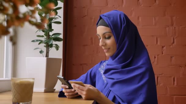 Vacker ung muslimsk kvinna i blå hijab omskrivning info från kort till hennes telefon, sitter i moderna café — Stockvideo