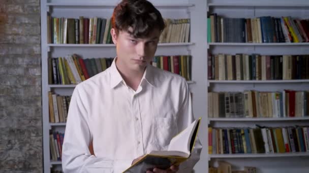 Sérieux brunette homme lit livre, regarder à la caméra, bibliothèque en arrière-plan — Video