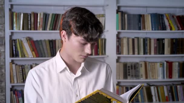 Hombre morena está leyendo libro, mirando a la cámara, biblioteca en el fondo — Vídeos de Stock