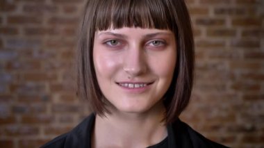 Kısa saç kesimi ve kameraya bakarak ve gülümseyen izole tuğla zemin üzerine yeşil gözlü genç kadın