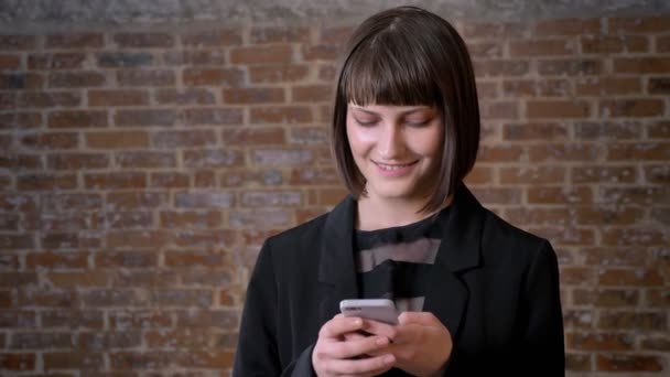 Young pretty woman with short haircut typing on phone and smiling, standing near brick wall — Stock Video