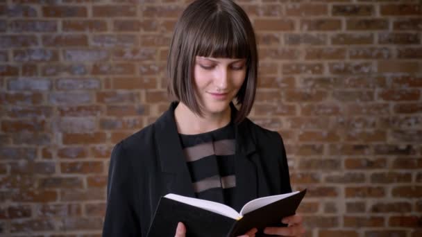 Jovem mulher concentrada com corte de cabelo curto leitura livro e de pé perto da parede de tijolo — Vídeo de Stock