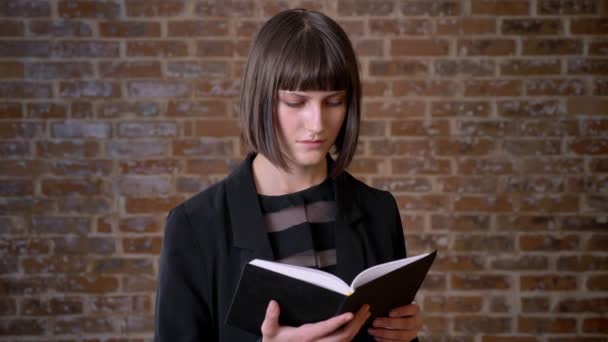 Giovane donna con taglio di capelli corto libro di lettura e sorridente alla macchina fotografica, isolato su sfondo di mattoni — Video Stock