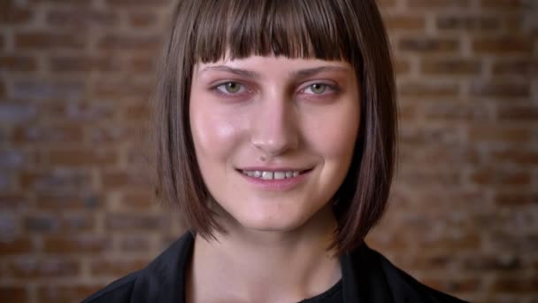 Jeune femme avec une coupe de cheveux courte et les yeux verts regardant la caméra et souriant, isolé sur fond de brique — Video