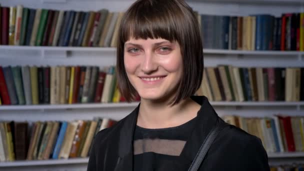 Junge glückliche, charmante Frau mit Kurzhaarschnitt, die in der Bibliothek steht, in die Kamera lächelt und lacht — Stockvideo