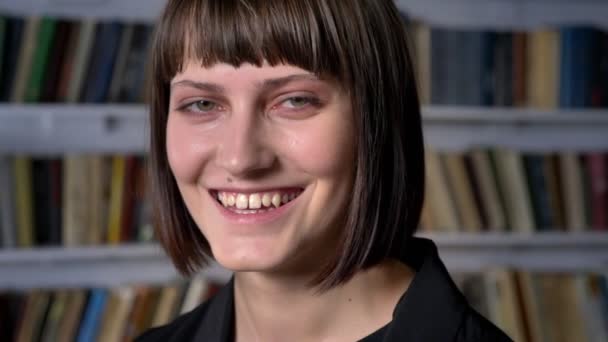 Ritratto di giovane bella donna in piedi in biblioteca e sorridente alla macchina fotografica, felice e allegra — Video Stock