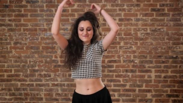 Beautiful woman with dark curly hair is watching at camera, dancing, smiling, jumping, brick background — Stock Video