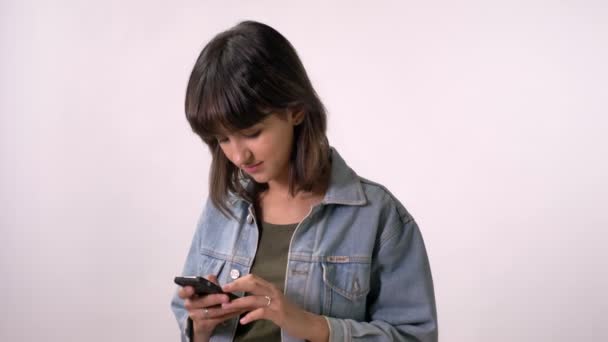 Jonge brunette meisje is bericht aan het typen op smartphone, kijken in de camera, glimlachen, witte achtergrond — Stockvideo