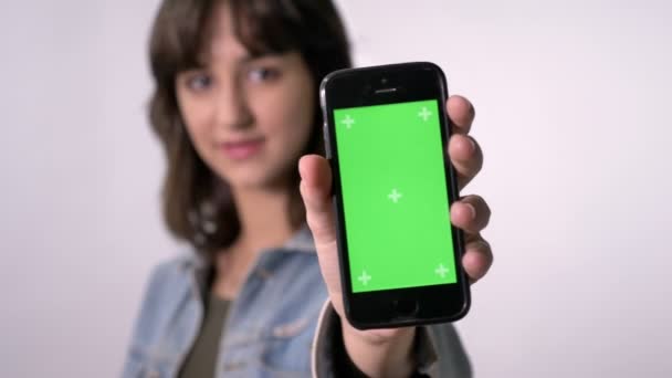 Junges brünettes Mädchen zeigt grünen Bildschirm des Smartphones, vor der Kamera, Kommunikationskonzept, weißer Hintergrund — Stockvideo