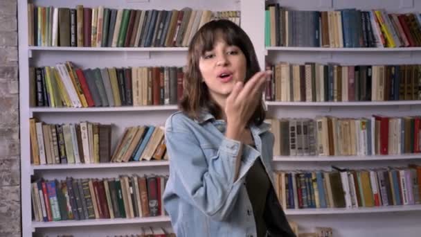 Young brunette girl is watching at camera, blowing a kiss, library on background — Stock Video