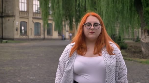 Gordura gengibre menina com óculos está de pé fora no parque em clima sombrio, assistindo a câmera — Vídeo de Stock