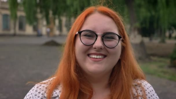 Dickes Ingwermädchen mit Brille steht bei trübem Wetter draußen im Park, guckt in die Kamera, kichert — Stockvideo