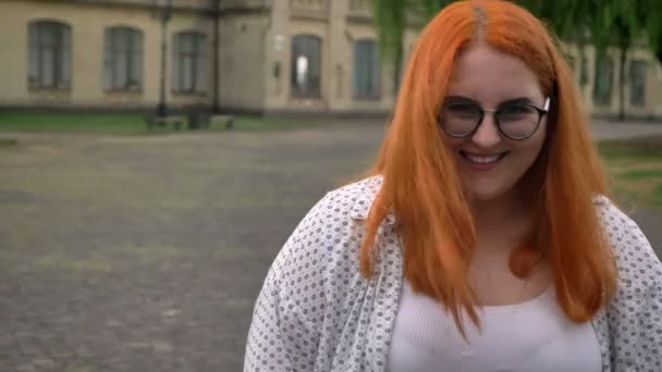 Fette Ingwermädchen mit Brille steht draußen im Park bei trübem Wetter, schaut in die Kamera, zeigt wie ein Zeichen — Stockvideo