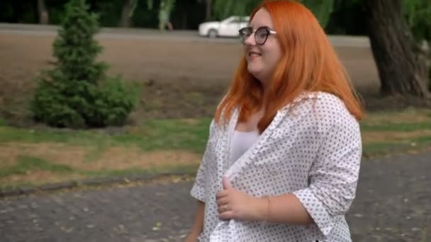 Жирная рыжая девушка в очках гуляет в парке в мрачную погоду, улыбается, боковой вид — стоковое видео