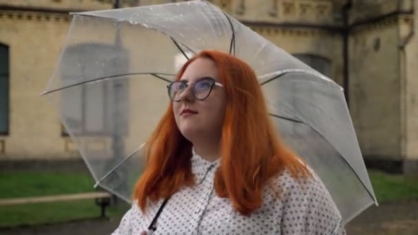 Жирная рыжая девушка в очках гуляет в парке в дождливую погоду, держит зонтик, вид сбоку — стоковое видео
