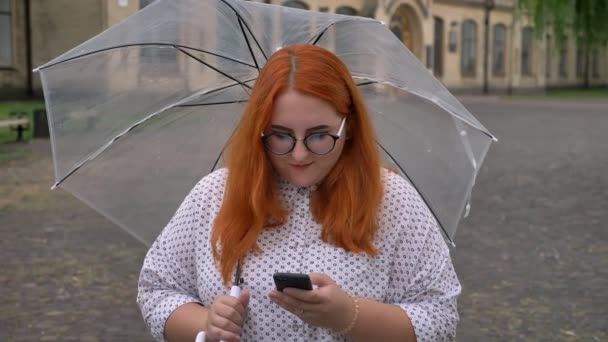 Tlusťoška zázvor s brýlemi je psaní zpráv na smartphone v parku v deštivém počasí, usmívající se, drží deštník — Stock video
