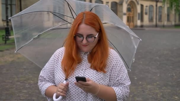 Chica gorda jengibre con gafas está escribiendo mensaje en el teléfono inteligente en el parque en tiempo lluvioso, riendo, sosteniendo paraguas, concepto de comunicación — Vídeos de Stock