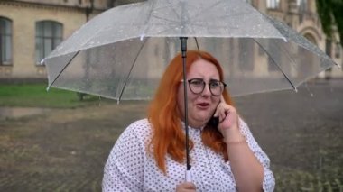 Gözlüklü şişman zencefil kız konuşurken telefon, holding şemsiye, iletişim kavramı üzerinde Park, yağmur altında duruyor