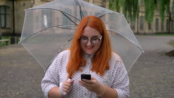 Fat ginger girl with glasses is typing message on smartphone in park in rainy weather, watching at camera, smiling, holding umbrella, communication concept — Stock Video