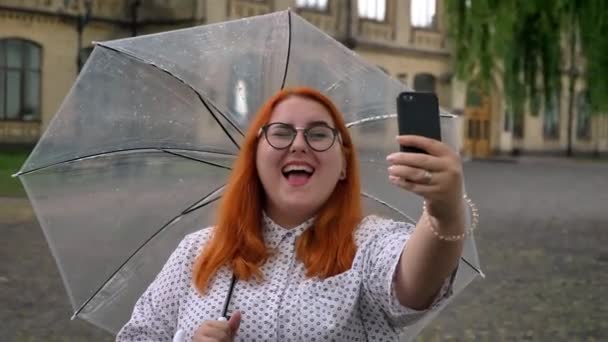 Dickes Ingwermädchen mit Brille macht Selfie auf Smartphone im Park bei Regenwetter, zwinkert, hält Regenschirm, Kommunikationskonzept — Stockvideo