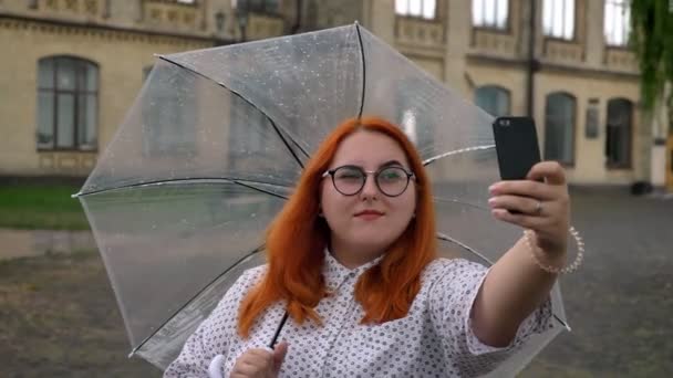 Жирная рыжая девушка в очках делает селфи на смартфоне в парке в дождливую погоду, улыбается, держит зонтик, коммуникационная концепция — стоковое видео
