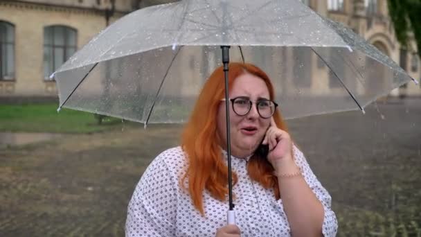 Gordura gengibre menina com óculos está de pé sob chuva no parque, falando no telefone, segurando guarda-chuva, conceito de comunicação — Vídeo de Stock