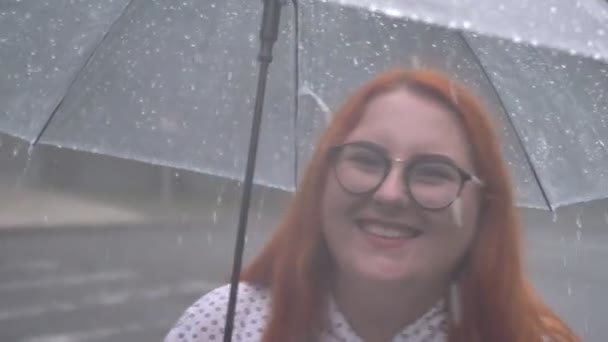 Жирная рыжая девушка в очках гуляет в парке под дождем, держит зонтик, смотрит в камеру, улыбается — стоковое видео