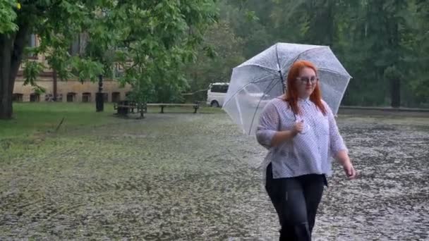Толстая рыжая девушка в очках гуляет в парке под дождем, смотрит вверх, держа зонтик — стоковое видео