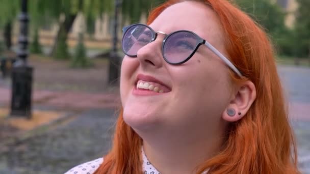 Chica gorda de jengibre con gafas está caminando en el parque bajo la lluvia, mirando en el cielo, sonriendo, concepto de inspiración — Vídeos de Stock