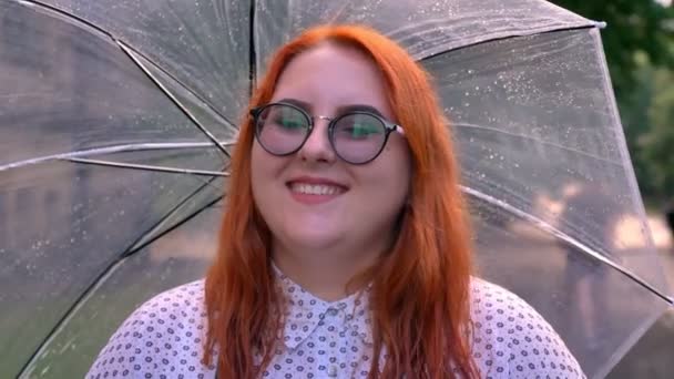 Жирная рыжая девушка в очках гуляет в парке в дождливую погоду, держа зонтик, улыбаясь, думая концепции — стоковое видео